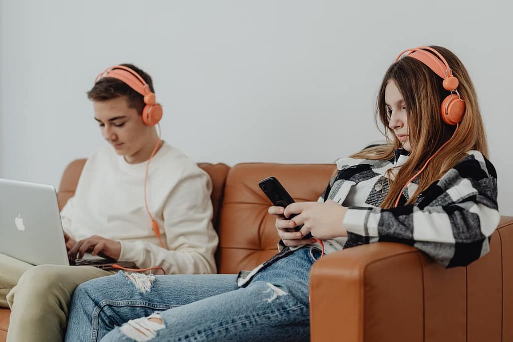 young girl using phone with headphones on, young boy using macbook with headphones on